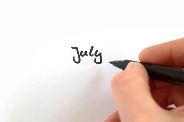 July, black handwritten word on white paper, hand holding pen