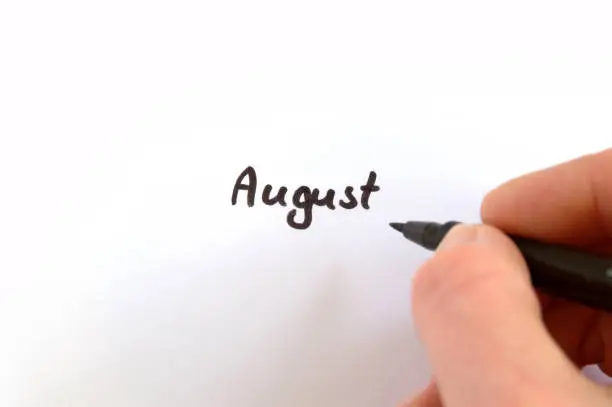 August, black handwritten word on white paper, hand holding pen