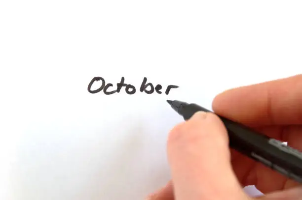 October, black handwritten word on white paper, hand holding pen