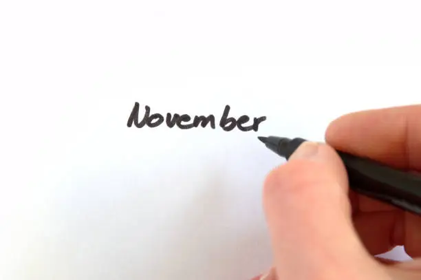 November, black handwritten word on white paper, hand holding pen