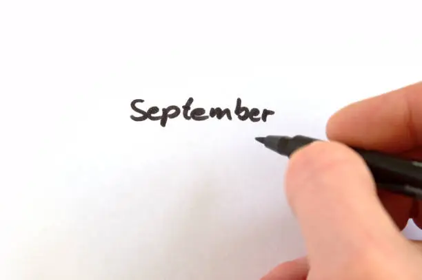 September, black handwritten word on white paper, hand holding pen