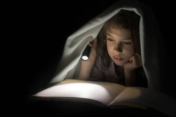 istock Girl reading a book with a flashlight in the dark under a blanket. 957438950