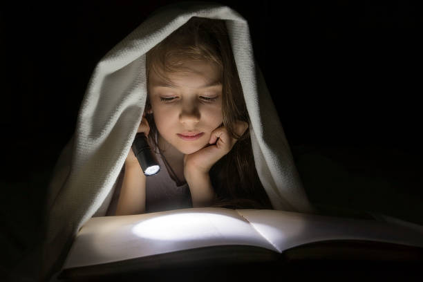 istock Girl reading a book with a flashlight in the dark under a blanket. 957439080
