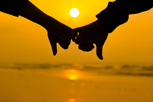 couple holding each other hands using little pinky finger in background of sunrise at the beach