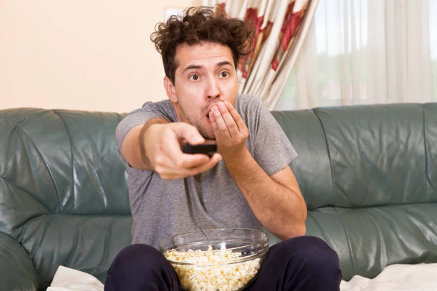istock Excited young man with the remote and popcorn on the couch watching tv 957539934