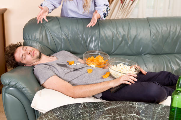 angry wife and sleeping husband on the sofa and junk food - routine foods and drinks clothing household equipment imagens e fotografias de stock