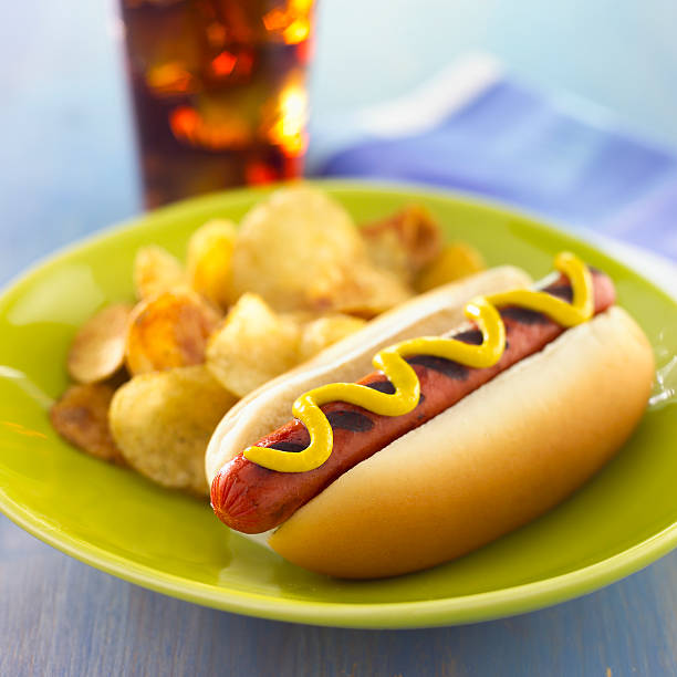 Traditional ballpark hotdog with mustard message, ketchup, onions, relish in bun on a weathered wood picnic table.  Served on a sanitary white paper fluted hot dog tray  with baseball to complete the theme.