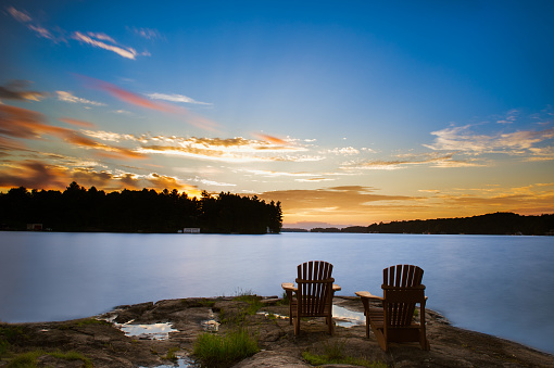 Two Adirondack chairs sitting on a rock facing a blue calm lake at sunset.