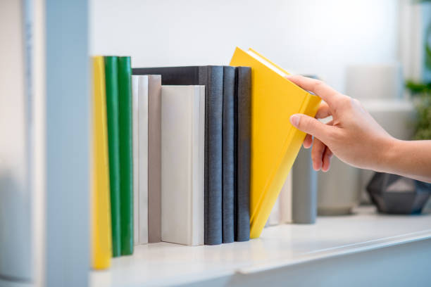 istock Male hand choosing and picking white book on white bookshelf in public library 957669632