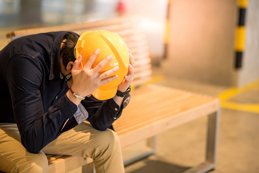 Young Asian engineer or architect man feeling tried and worried while sitting on bench holding his personal yellow protective safety helmet. depression from hard work concept