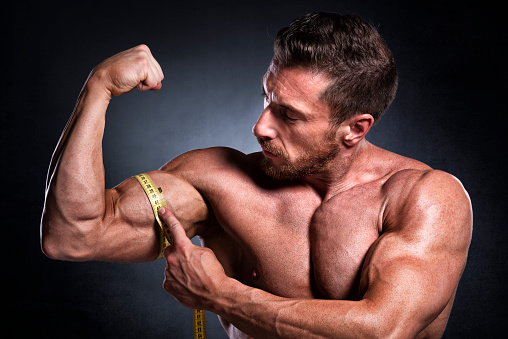 Studio shot of male fitness athlete while measuring bicep with tape measure.