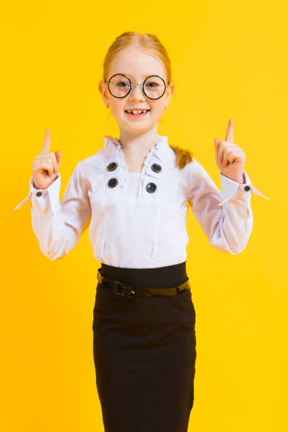 Girl with red hair on a yellow background. A charming girl in transparent glasses points her fingers up. Portrait of a beautiful girl in a white blouse and black skirt. see through leggings stock pictures, royalty-free photos & images