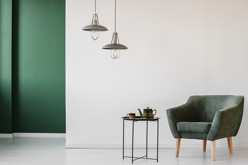 Upholstered dark armchair and an industrial side table with a tea kettle and cup in a minimalist living room interior with a green corner