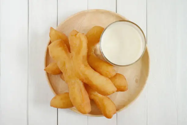 Photo of Pa Tong Go, Thai style Chinese crullers, with a glass of soy milk in wooden plate on white wooden background. Top view.