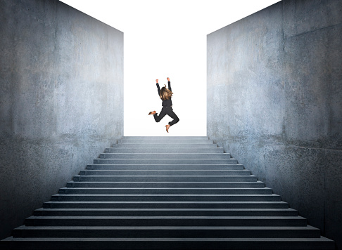 A businesswoman who has reached the top of the stairs throws up her arms and kicks out her legs as she jumps for joy having reached the top.