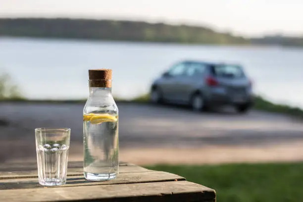 A bottle of water with lemon and a glass. Table by the lake. In the background a car in the parking lot. Season of the spring.