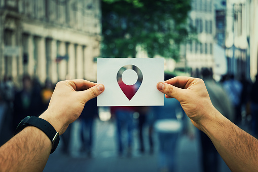 Close up of man hands holding a white paper sheet with a pointer icon inside. GPS  searching for someone on a crowded city street. Travel navigation and planning location route.