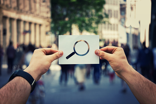 Close up of man hands holding a white paper sheet with magnifier icon inside. Searching for someone on a crowded city street background.