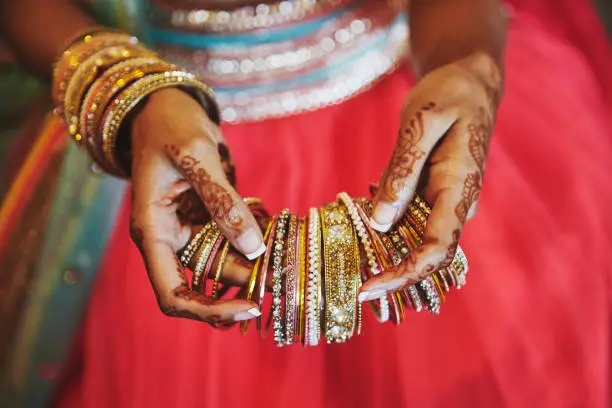 Photo of The hand with mehndi of Indian bride holding a lot of glitter bracelets (bangle) with red legenha background, close-up