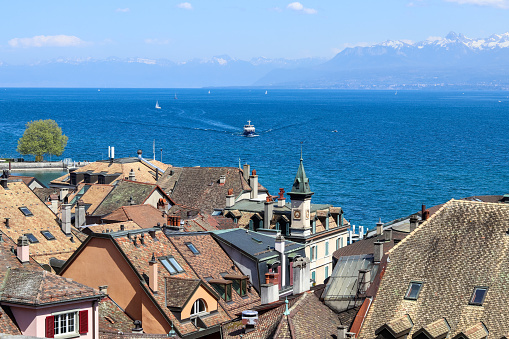 Amazing view over the roofs of old Nyon town at blue Geneva lake or Lac Leman with sailboats and ship and mountains with snow tops at background, Switzerland, Europe