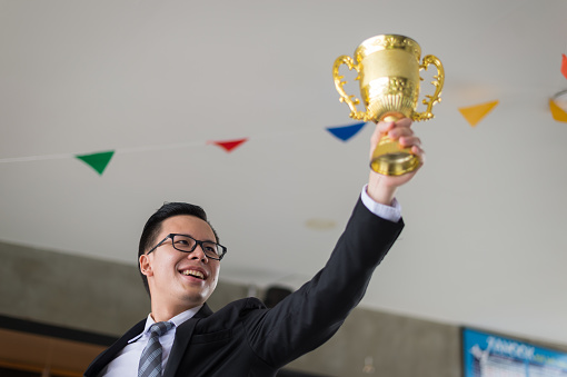 Asian businessman raising up hand and holding a golden trophy cup to cheerful and celebrated his successful in career and mission. concept of winner and competition.
