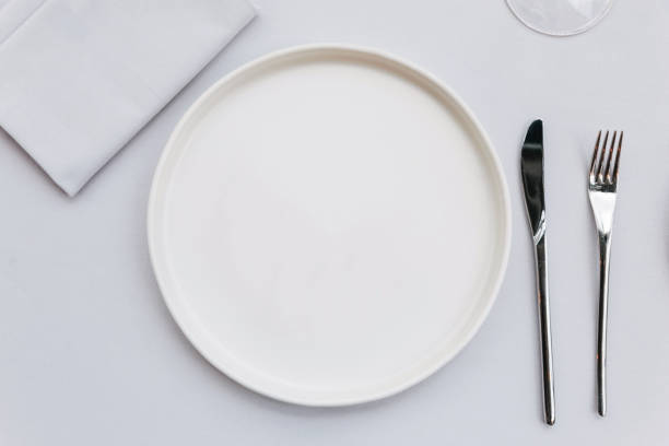 istock Top view of white empty plate on white tablecloth with knife, fork, napkin and wine glass. 957994588