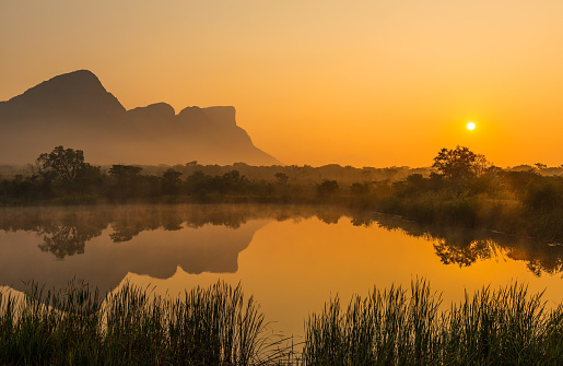 Sunrise inside the Entabeni Safari Game Reserve with a reflection of the Hanging Lip of Hanglip mountain peak in a misty swamp lake located near Kruger Park, Limpopo Province, South Africa.