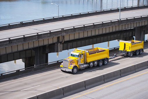 Big rig powerful classic yellow tipper semi truck with two dumps for carry more cargo running on overpass intersection road along the river and another elevated road on the side