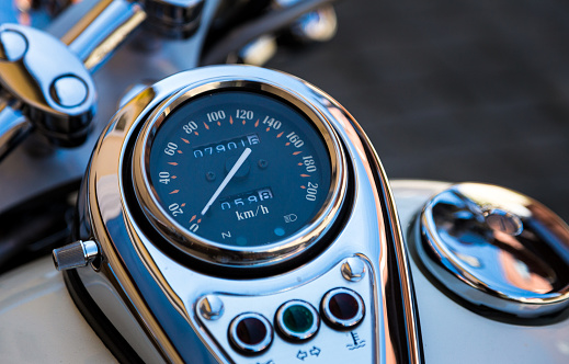 Close up color image depicting the chrome handlebars and large circular odometer of a vintage, classic motorbike. Focus is sharp on the speedometer, while the background of the shiny metal handlebars is blurred out of focus. Room for copy space.