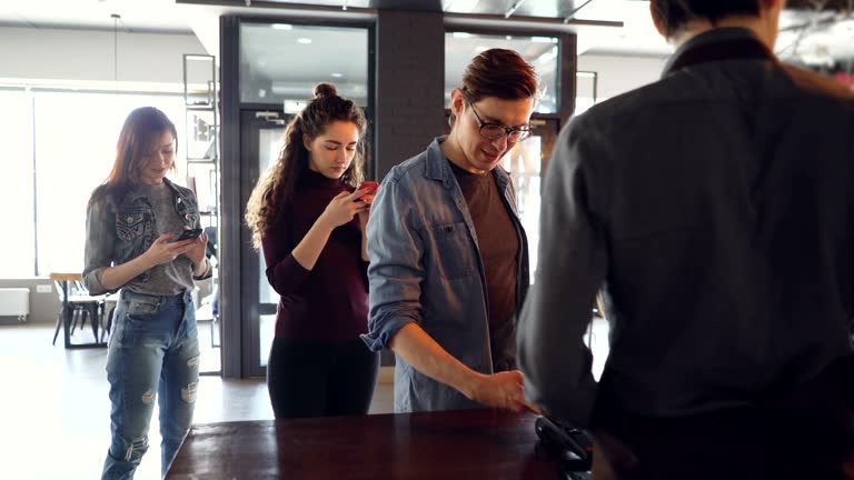 Cheerful customer in cafe is making payment with smartphone while queue of clients are waiting and using mobile phones. Takeaway drinks and modern banking concept.