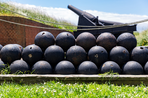 A stack of historic cannon balls stand at the ready in front of large cannons in firing position.