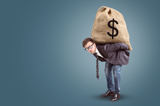 A businessman with a straining expression is carrying a large bag with a dollar sign on it.