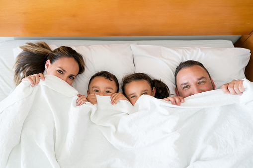 Beautiful latin american family relaxing in bed covering their mouths with a bedsheet all looking at camera very playfully