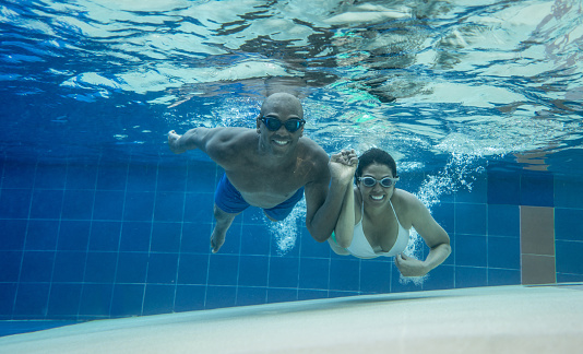 Young diverse couple having fun swimming under water looking at camera smiling very happy while holding hands