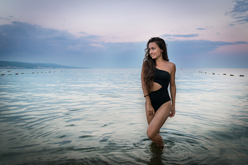 Young shapely woman stands in sea on background of colorful sunset and calm seascape. Pretty girl in swimwear went into the water and looks away.