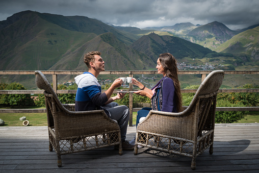 Young couple sits together on the terrace with cups of coffee in the morning. Man sends air kiss to girlfriend. Travelers having breakfast with beautiful view.