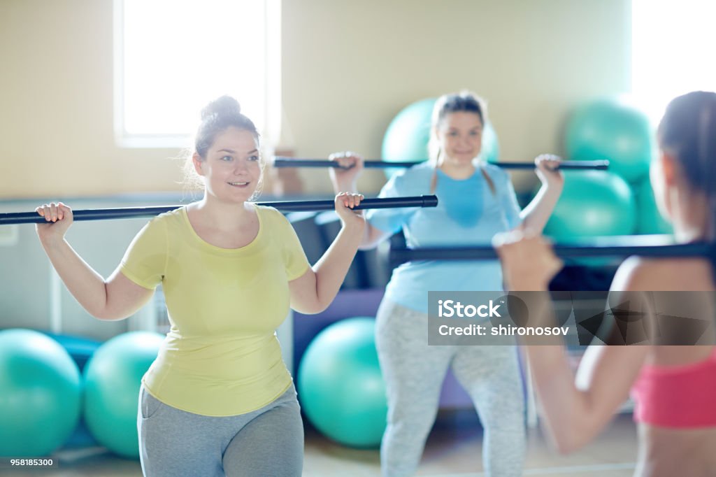 Repeating after instructor Two happy oversized women with gymnastic bars doing exercises after their instructor in gym Overweight Stock Photo