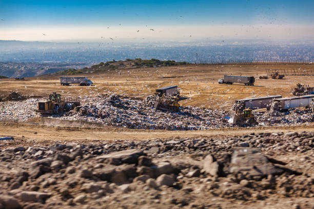 Shot of a truck dumping more garbage onto a U.K. landfill site alongside a bulldozer. Residential area visible in far background. Dust from rubbish in air. Black smoke from moving bulldozer is visible. Plastic bin bags, cardboard and wood visible amongst the garbage. 