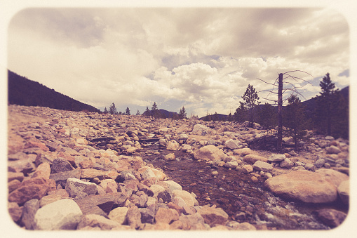 View from Rocky Mountain National Park in Colorado with stream over rock landscape, mountains and ominous sky.  Image has a vintage tone filter and film effect