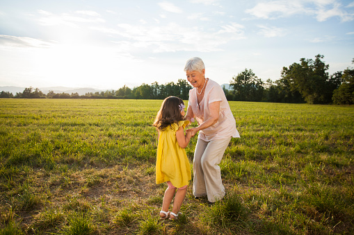Female senior adult holding hands with small girl, her grandchild and dancing in meadow. Nice sunny summer day.