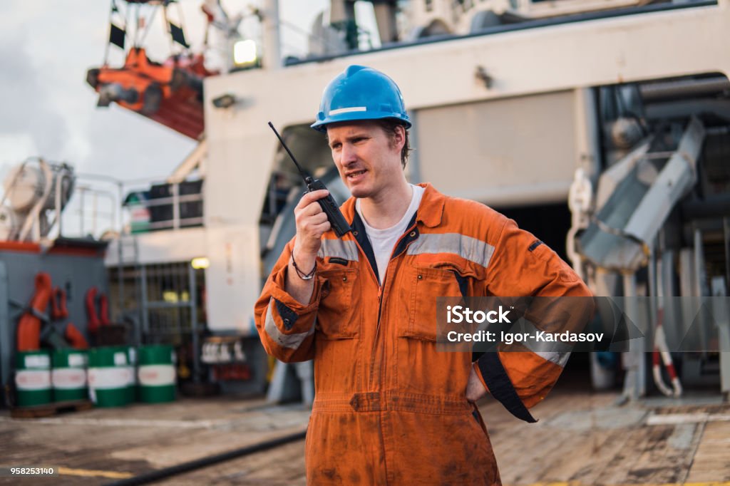 Marine Deck Officer or Chief mate on deck of ship with VHF radio Marine Deck Officer or Chief mate on deck of offshore vessel or ship , wearing PPE personal protective equipment - helmet, coverall. He holds VHF walkie-talkie radio in hands. Ship Stock Photo