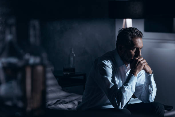 retrato de un hombre maduro solitario con sentado en una cama en una habitación gris con botellas de pie de alcohol alrededor de la depresión - assistance help dependency business fotografías e imágenes de stock