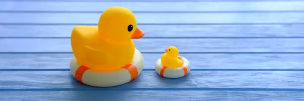 Photo of Large yellow rubber duck looking down on a small yellow rubber duck, both on inflatable life rings, scene set on a blue wood background which, conceptually represents water.