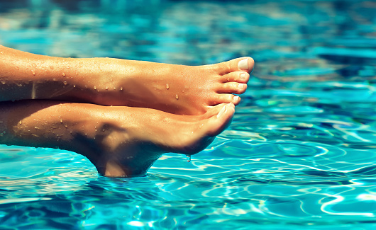Tanned, well-groomed crossed woman's feet, covered by drops of clean water is resting above blue, moving surface of waterpool.Pedicure, feet care and Spa. Simbolik image of comfortable rest in vacation.