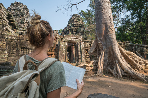 Young woman traveling in Cambodia visiting the temples of Angkor wat complex and reading guidebook. People travel discovery Asia concept. Shot at sunset, one woman only, adventure and exploration in Siem Reap, Southeast Asia.