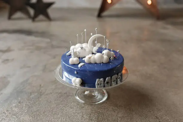 Gourmet blue birthday cake with white decor and candle number one on glass stand in loft interior. Home indoors cake-smash first year concept.
