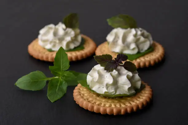 Bisquit cracker appetizers with cream cheese and basil topping on black stone background