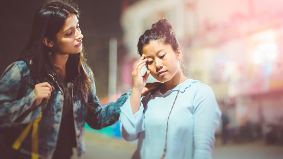 Asian, Indian multi ethnic female friend standing together on city street in evening and one friends is consoling other who is  sad.