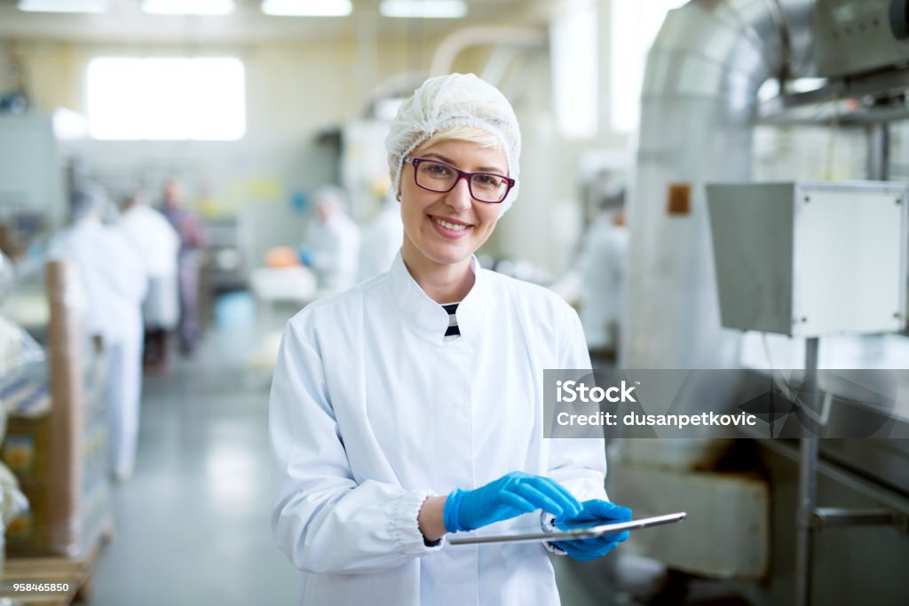 若いうれしそうな美しい女性労働者滅菌布タブレットを保持していると、工場の生産ラインに近いカメラに笑顔で。 - 食べ物のロイヤリティフリーストックフォト
