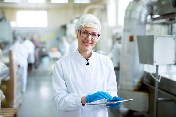 young joyful beautiful female worker in sterile cloths holding a tablet and smiling for the camera near factory production line. - food hygiene imagens e fotografias de stock